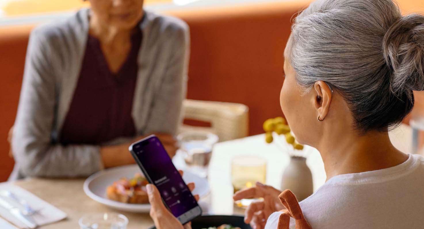 Woman sitting at a cafe controlling her hearing aids with the my starkey app.